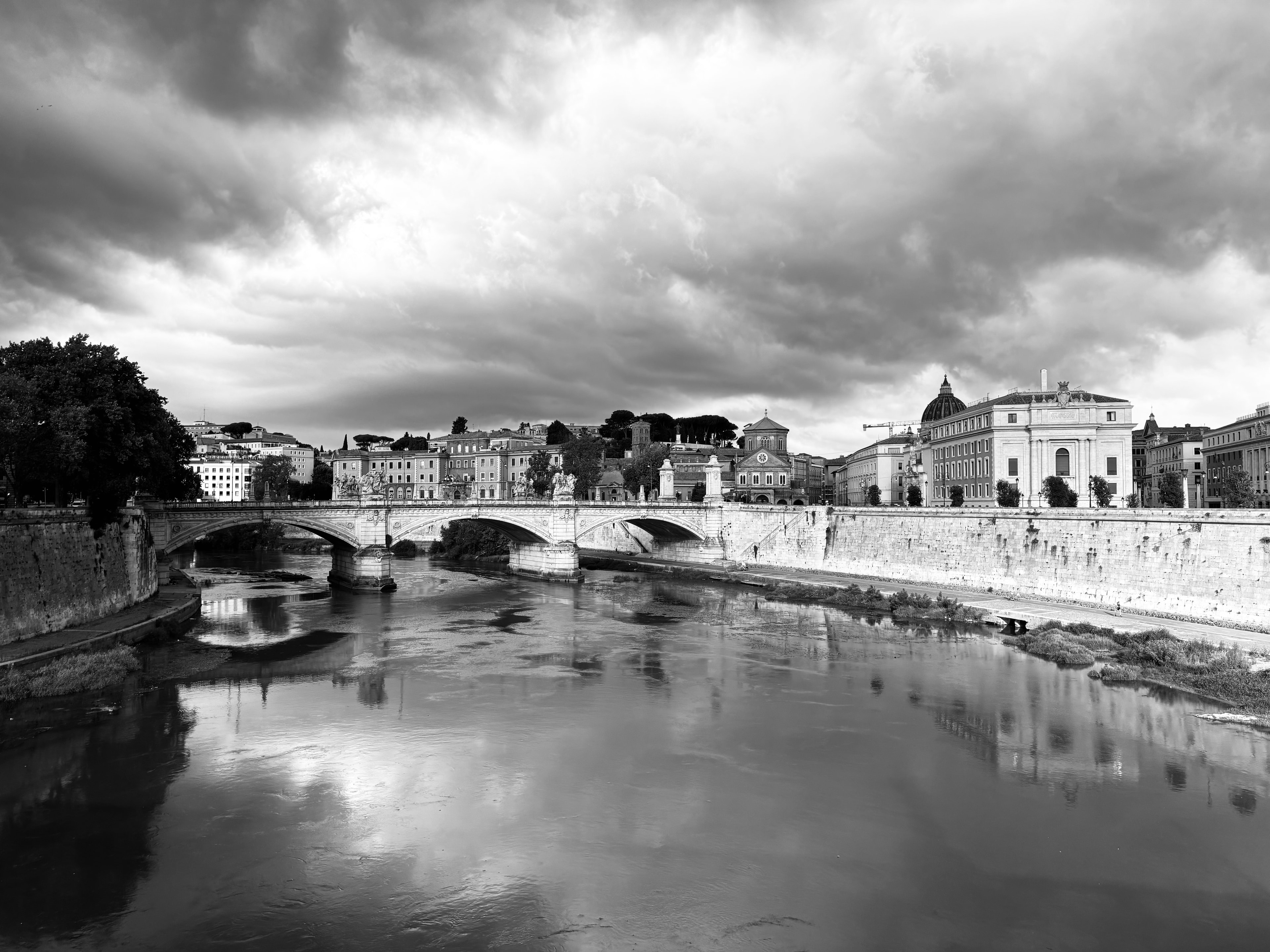 Black and white riverside cityscape with bridge and dramatic storm clouds